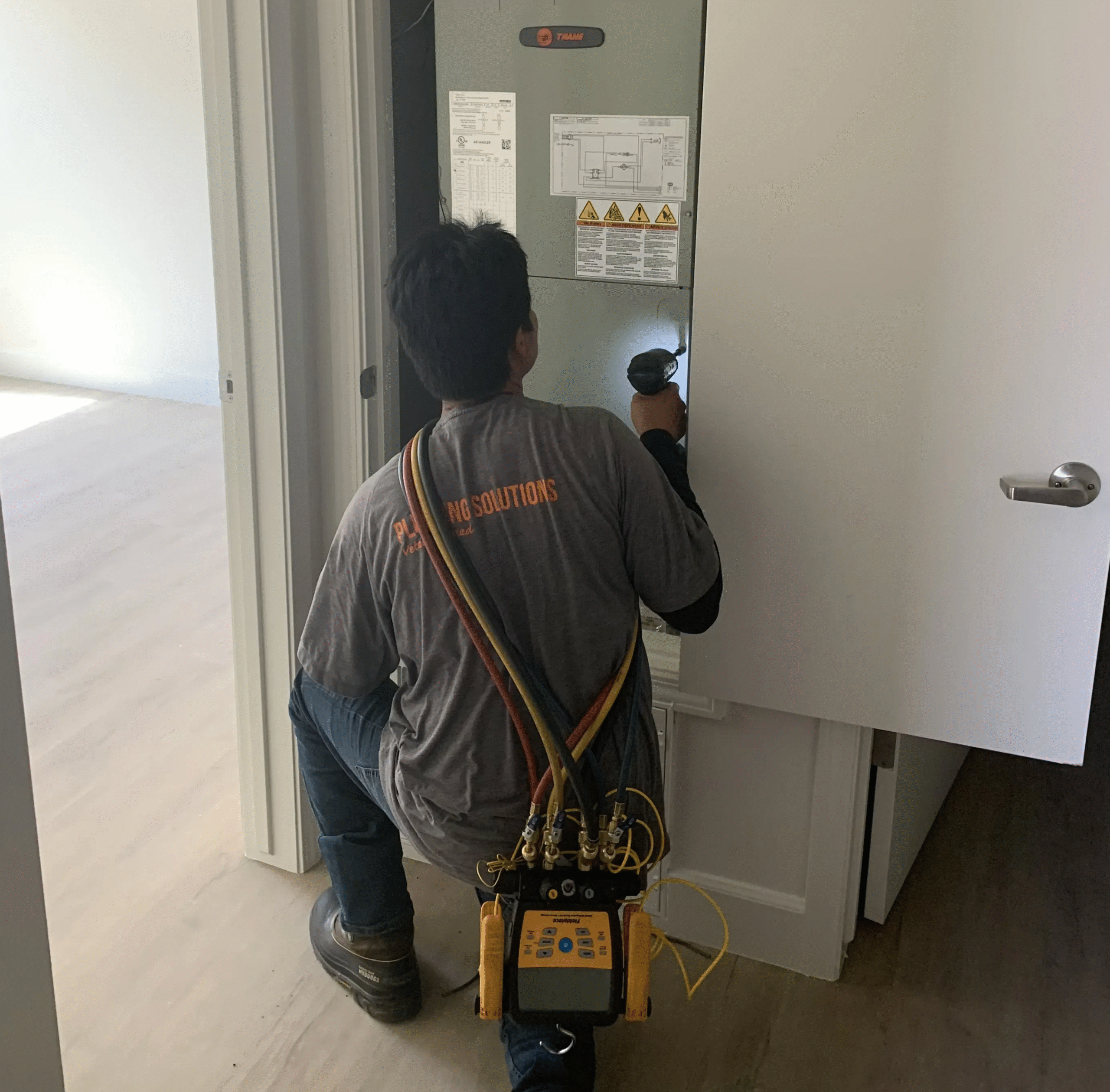 A technician kneels in front of an electrical panel, inspecting it with a flashlight. He wears a gray shirt and has a diagnostic device strapped over his shoulder, ensuring top-notch furnace maintenance for Central Valley homes.