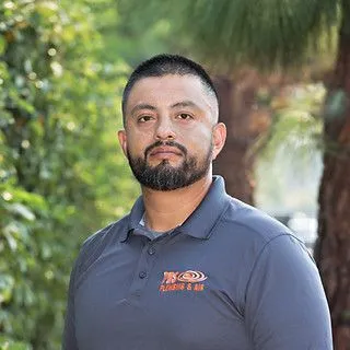 Man with a beard and short hair stands outdoors wearing a gray PDS Plumbing polo shirt with an orange logo, with trees and greenery in the background.