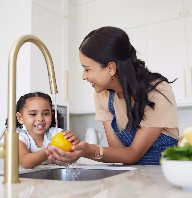 An adult and a young child smile while washing a yellow bell pepper under a kitchen faucet, with vegetables on the counter—a moment made possible by reliable plumbing services.