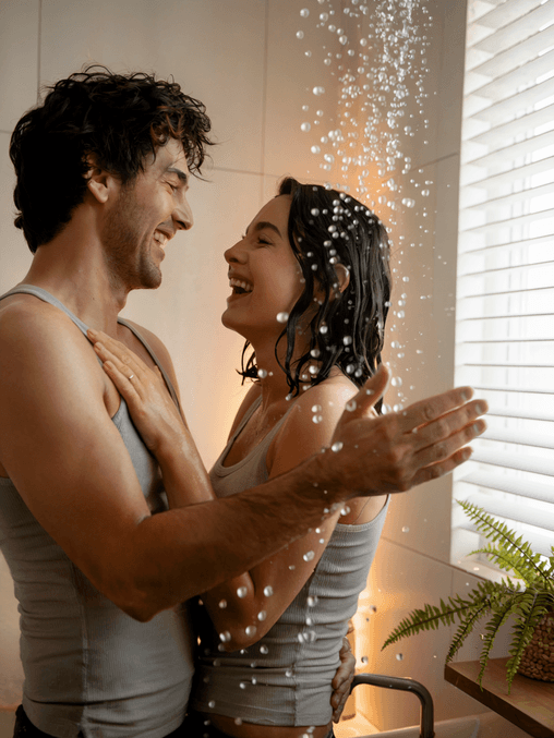 Two people wearing gray tank tops stand close together, smiling and laughing under a shower, enjoying the warm spray from their new water heater in a bathroom with a window and a potted fern visible.