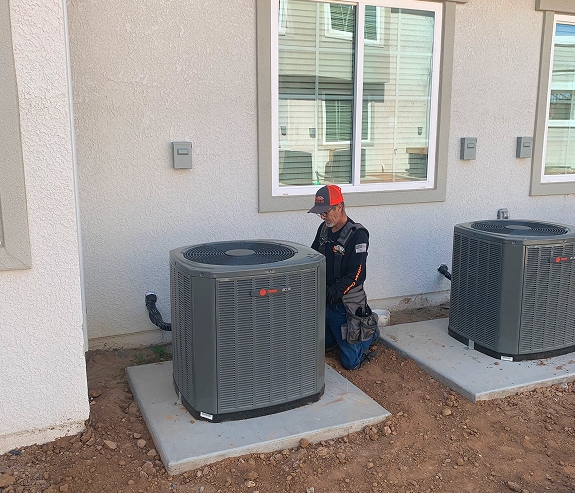 A technician kneels next to an outdoor air conditioning unit outside a building, performing maintenance or inspection work to help ensure expert indoor air quality.